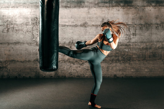 Dedicated strong brunette with ponytail, in sportswear, bare foot and with boxing gloves kicking sack in gym.