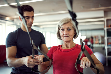A senior woman in gym with a young trainer doing exercise with TRX.
