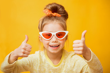 Surprised funny little girl in sunglasses shows thumbs up and looking camera over yellow
