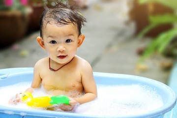 A happy baby child take a shower with a duck toys. She sitting in a bathtub with many bubbles soap. Infant girl aged of 1 years looking at a camera.