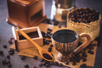 Brew black coffee in coconut cup and morning lighting. Roasted coffee beans in a bamboo basket and wooden spoon. Vintage coffee grinder and pot. Concept of coffee time in morning or start the new day.