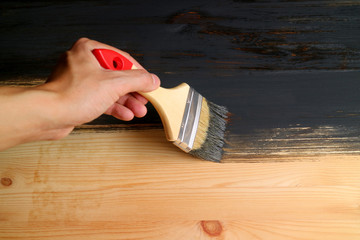 Man's hand holding brush painting wood plank with dark grey paint