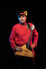 A brunette man in a folk shirt plays a balalaika in scenic blue and red light on a black stage