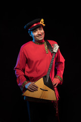 A brunette man in a folk shirt plays a balalaika in scenic blue and red light on a black stage