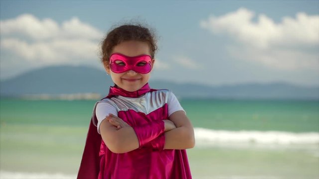 Beautiful Little Girl in the Superhero Costume, Dressed in a Pink Cloak and the Mask of the Hero. Plays on the Background Sea and Blue Sky and Clouds. Concept of a Happy Childhood.