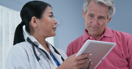 Japanese woman primary care doctor using tablet computer while explaining treatment to senior Caucasian male patient. Low-angle view of physician talking to aging man