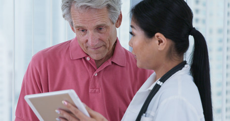 Obraz premium Elderly Caucasian male patient asking Japanese woman doctor questions while looking at tablet computer together. Female primary care physician and her patient discussing treatment