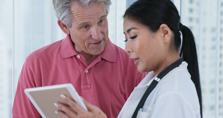 Obraz premium Elderly Caucasian male patient asking Japanese woman doctor questions while looking at tablet computer together. Female primary care physician and her patient discussing treatment