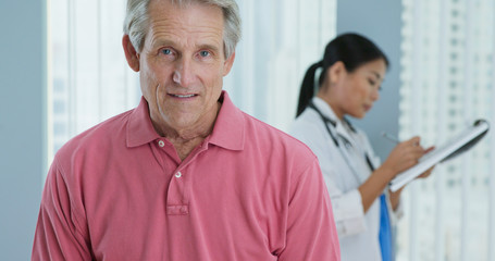 Fototapeta premium Portrait of senior Caucasian male patient looking at camera and smiling while doctor works in background. Attractive older man in hospital with female physician out of focus behind him