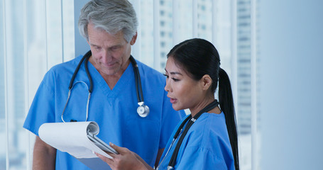 Medium shot of Japanese woman doctor and Caucasian male nurse going over paperwork in hospital. Two friendly medical professionals wearing blue scrubs working together.