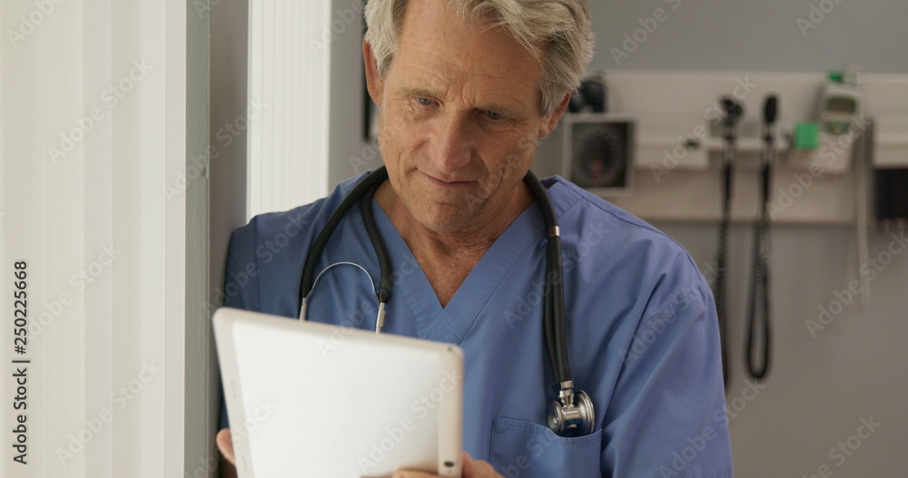 Wall mural Happy Senior Caucasian male doctor reading tablet computer with medical history of patient and smiling. Older man medical professional looking over positive results on pad