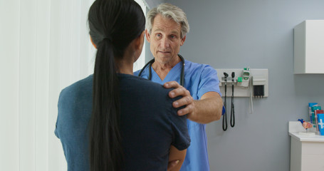 Medium shot of supportive senior Caucasian male doctor listening to female patient with his hand on her shoulder in hospital exam room. Woman talking to her nurse or OBGYN