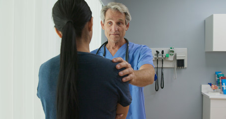 Obraz premium Medium shot of supportive senior Caucasian male doctor listening to female patient with his hand on her shoulder in hospital exam room. Woman talking to her nurse or OBGYN
