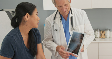 Asian woman sitting on exam table looking at tablet computer with knee x-ray as doctor explains treatment of injury. Senior medical professional using device with patient