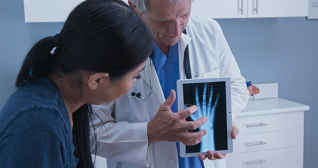 Doctor using tablet computer with x-ray of hand to talk about arthritis with young patient. Senior Caucasian male medical professional with screen demonstrating joint injury to woman - obrazy, fototapety, plakaty