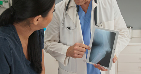 Close up on hands of doctor explaining knee injury while holding tablet computer with x-ray. Patient watching medical professional demonstrate problem with her joints
