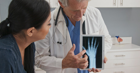 Doctor using tablet computer with x-ray of hand to talk about recovery with female patient. Senior Caucasian male medical professional with screen demonstrating arthritis to woman