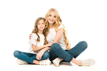 mother and daughter embracing while sitting on floor with crossed legs and looking at camera on white background