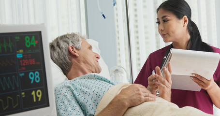 Asian woman doctor talking with senior Caucasian male patient lying in hospital bed. Medical professional monitoring symptoms of ill patient