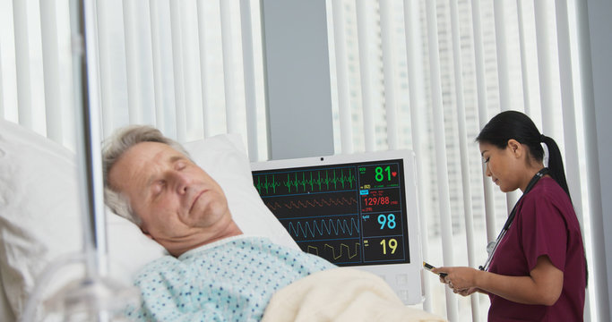 Doctor Standing In Window Of Hospital Room Text Messaging Family On Mobile Phone While Patient Recovers In Bed. Senior Man In Foreground With Surgeon Updating Loved Ones In Background