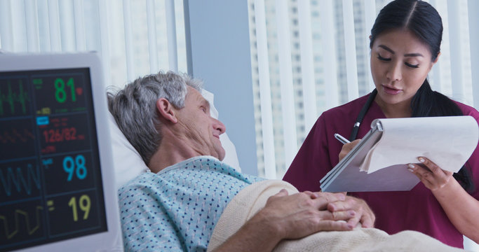 Asian Woman Doctor Talking With Senior Caucasian Male Patient Lying In Hospital Bed While Nurse Moves In Foreground. Medical Professional Monitoring Symptoms Of Ill Patient