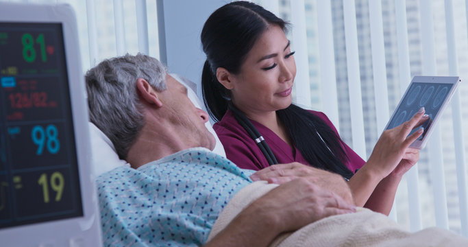 Senior Caucasian Male Patient In Hospital Bed Asking Questions About Brain Scans To Japanese Woman Doctor. Medical Professional Showing Medical Images To Older Man On Tablet Screen