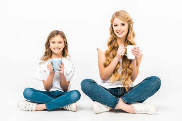 smiling mother and daughter holding coffee cups while sitting on floor with crossed legs and looking at camera on white background