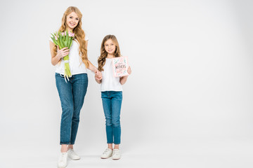 Fototapeta premium smiling mother with bouquet of pink tulips, and smiling daughter with happy mothers day greeting card holding hands while looking at camera on white background