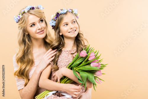 beautiful mother and adorable daughter in floral wreaths looking away and smiling on yellow background