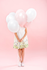 child in delicate white dress hiding behind white and pink air balloons on pink background