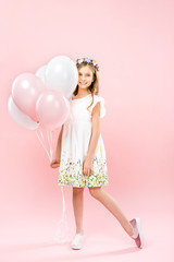 adorable child in elegant white dress and floral wreath holding festive air balloons on pink background