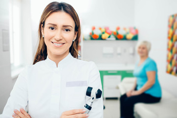 Doctor dermatologist with dermatoscope, smiling while looking at camera. Behind her sitting senior patient.