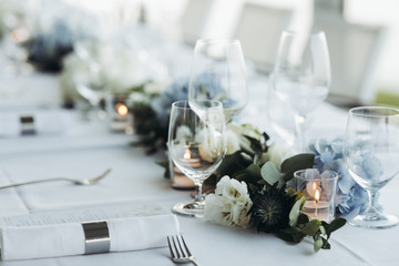 Wedding table decoration. Floral garland of greenery and blue flowers lies between glasses on the white table