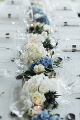 Wedding table decoration. Floral garland of greenery and blue flowers lies between glasses on the white table