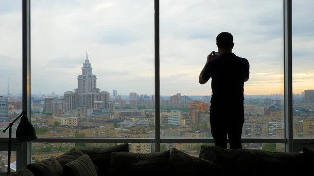 silhouette of a man trying to take a photo at the Apartment or office with panoramic windows and a beautiful view of the city of Moscow. Long shot.