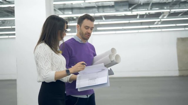 A woman and a man are walking along the hall with architectural drawings