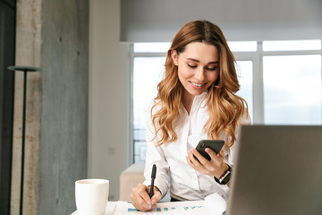 Woman dressed in formal clothes shirt indoors using mobile phone writing notes.