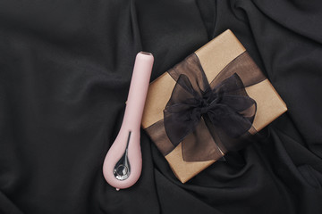 Sex toy gift for the women you love. Top view of pink vibrator for her and gift box with a bow on a black silk.