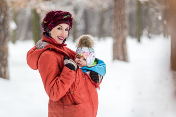 Mother carrying her baby girl wears red jacket and sling.