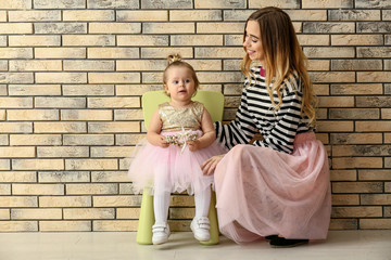 Young woman with her cute baby daughter dressed as little princess near brick wall