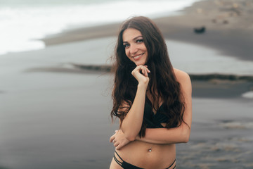 Portrait of happy girl with beautiful smile. Young woman in swimsuit posing on black sand beach