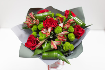 A bright bouquet of fresh flowers (Alstroemeria, Chrysanthemum, Rose, Aspidistra leaves. Primary colors: red and green) in a package made of gray paper in a vase on a light background.