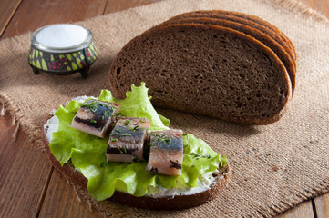 Sandwich with herring fillet, lettuce, dill and cream. Near dark rye bread and salt in salt shaker on burlap.