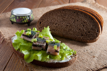 Sandwich with herring fillet, lettuce, dill, chives and cream. Near dark rye bread and salt in salt shaker on burlap.
