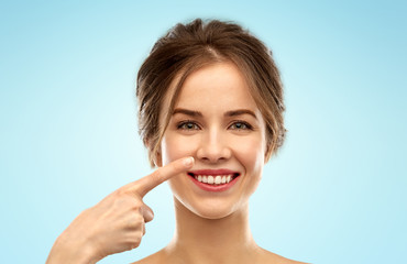 beauty, people and rhinoplasty concept - beautiful young woman pointing to her nose over blue background
