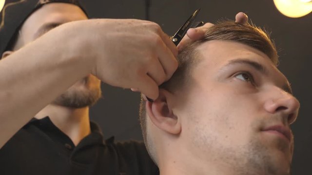Male barber combing and cutting hair of young customer by hairbrush and scissors in salon. Caucasian hairdresser trimming hair of his client in barbershop. Hairstyling process. Close up Slow motion