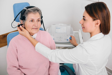 Senior patient wearing headphones, sitting near audiologist during ear exam at hearing clinic