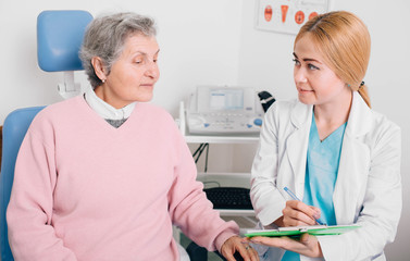 Fototapeta premium Friendly doctor consulting elderly patient in clinic and recording to medical chart