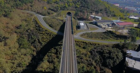 FLYING ABOVE THE TRAIN BRIDGE IN A GREEN VALLEY AND APPROACHING TO THE TUNNEL WITH FACTORIES, HOUSES AND THE CITY OF OURENSE