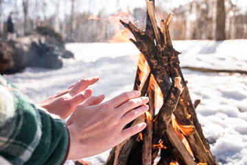 Basking near a campfire in a snowy birch forest. Female person getting warm near a fire on a sunny winter day in the woods - obrazy, fototapety, plakaty